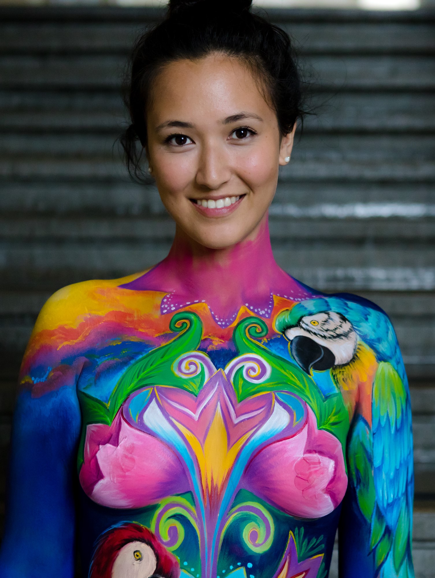 Photo Daniel Janesch WB Academy 2018 Smiling woman with elaborate bodypainting featuring vibrant tropical designs. Her chest and shoulders are painted in bright colors with pink flowers, green swirls, a colorful parrot on her right shoulder, and a red bird on her left side, blending into a sunset-like sky across her upper chest.